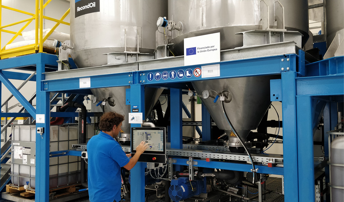 A male worker in a blue shirt operating a touchscreen control panel in an industrial setting. Behind him are large metal tanks supported by a blue metal framework, with signs indicating safety precautions and EU funding. The surrounding area includes various pipes, storage containers and additional equipment, all within a clean, organised environment.