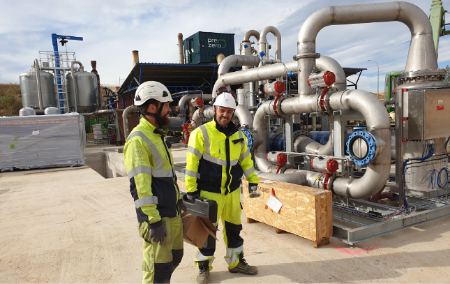 Two male workers in high-visibility safety gear stand in an outdoor industrial area, discussing equipment. They are surrounded by a complex network of large metal pipes, valves and industrial machinery. The background includes a building and various industrial structures.