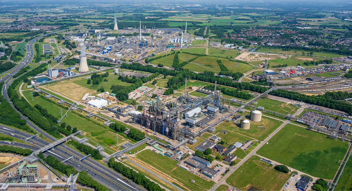 An aerial view of a large industrial complex with various buildings, storage tanks and pipelines, surrounded by greenery. There is a major highway running through the area, with bridges connecting different parts of the facility. The background features fields and a residential area, indicating a mix of industrial and rural land use.