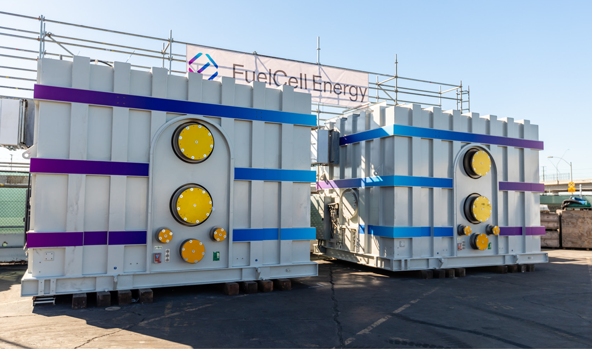 Two large, rectangular industrial units with prominent yellow circular fixtures on their fronts, marked with blue and purple horizontal stripes. These units are situated in an outdoor setting. The background includes metal scaffolding and a clear blue sky.