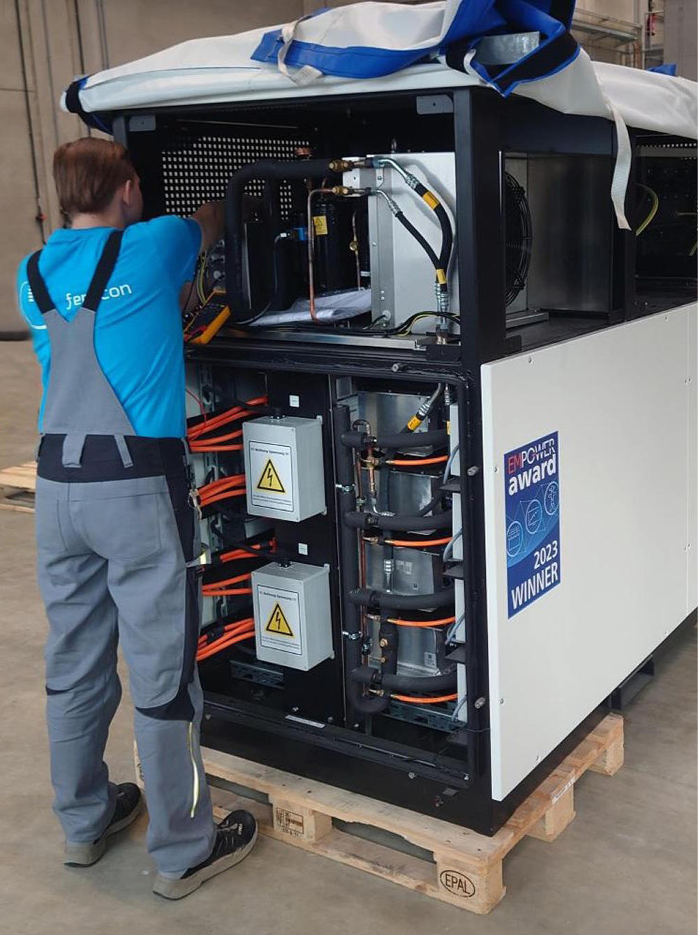 A technician in a blue shirt and grey overalls working on a large piece of equipment housed in a white and black casing. The equipment has several cables and warning signs, indicating high voltage. A sign on the equipment mentions an award, suggesting it is prize-winning technology.