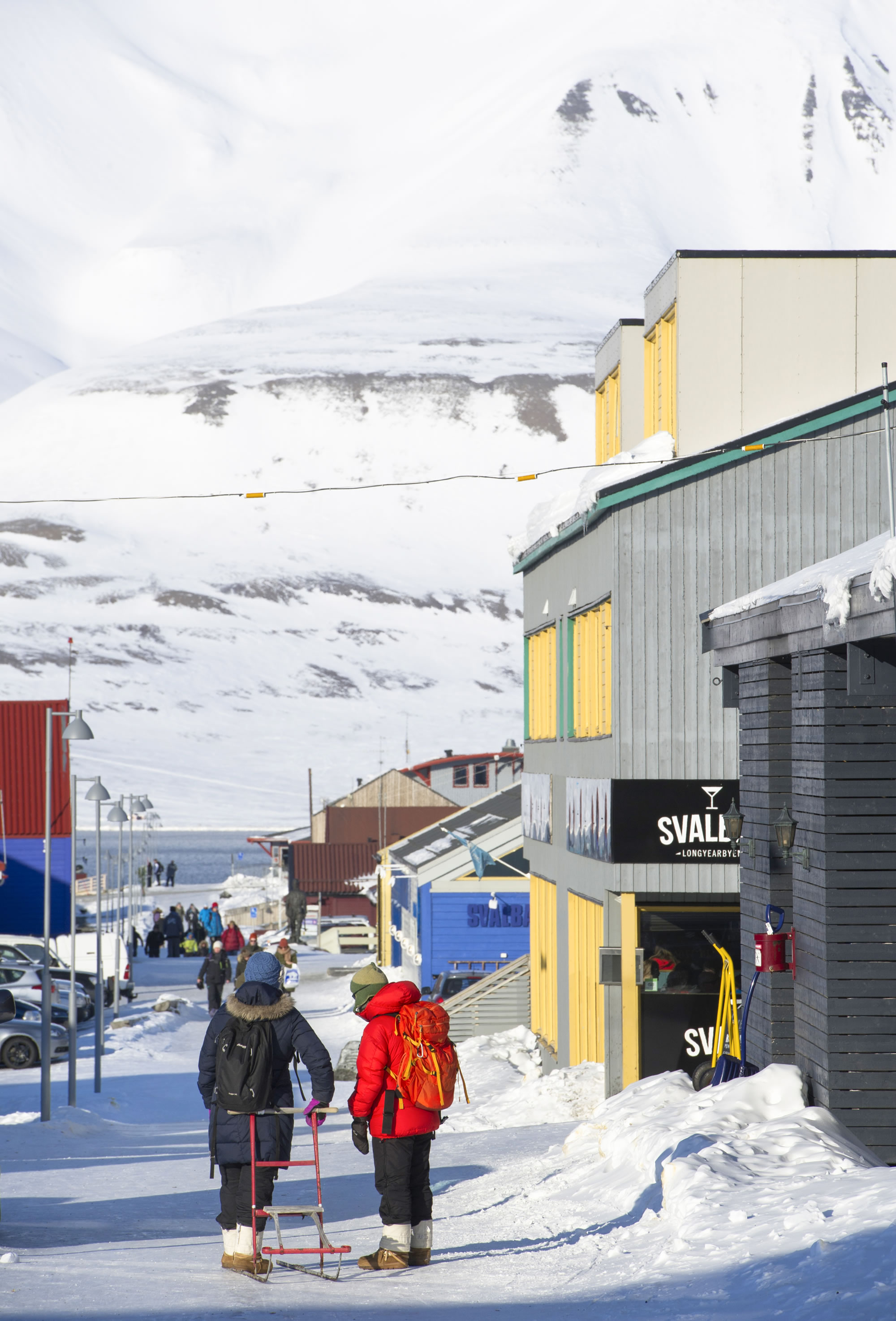 Deux personnes, en tenue d’hiver, marchent dans une rue enneigée bordée de maisons colorées, face à la mer, avec une montagne enneigée en arrière-plan.