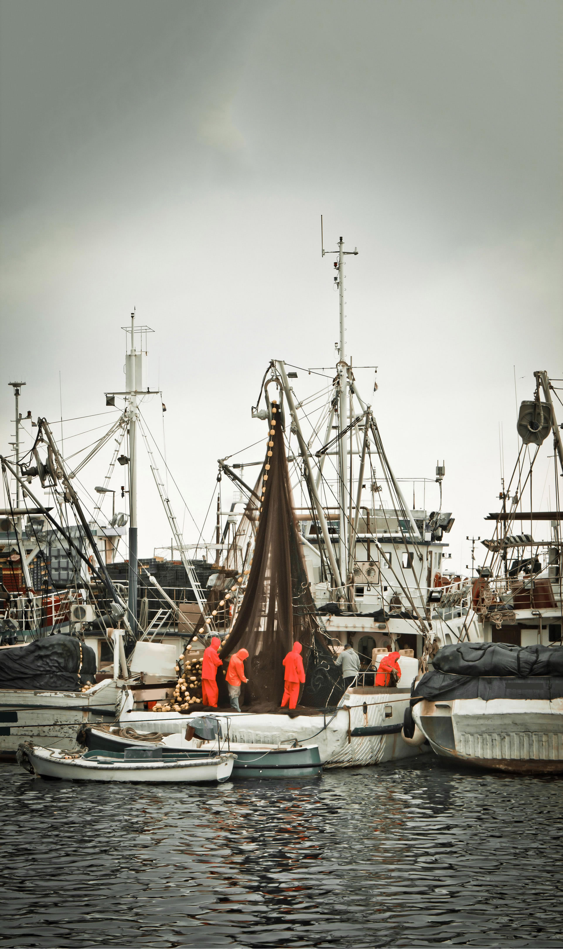 Pescari în echipament profesionist care manipulează o plasă de pescuit pe un trauler, în cadrul unei flote de ambarcațiuni într-un port de pescuit.