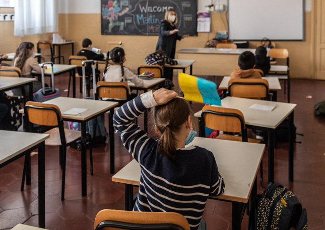 Diversi bambini accolgono in classe gli alunni ucraini sventolando bandierine ucraine.