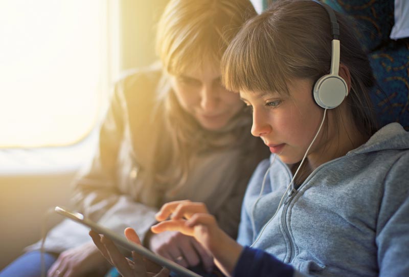 Dziewczynka w słuchawkach bawi się tabletem w pociągu, towarzyszy jej siedząca obok kobieta