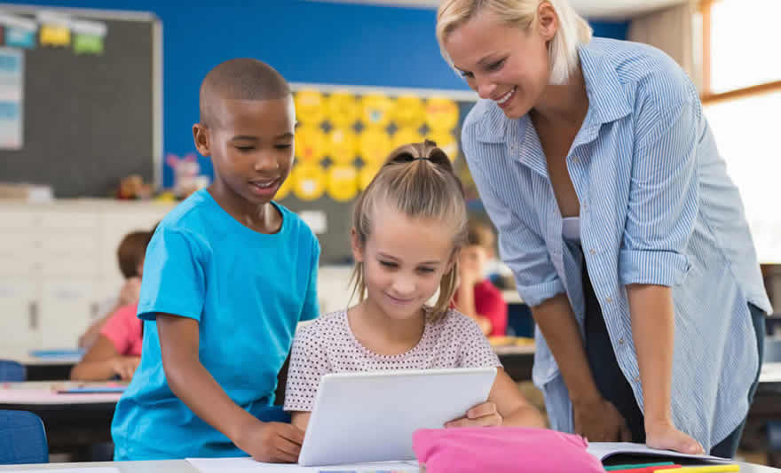 Image: A primary school teacher stands over two children as they collaborate on a tablet. © Fotolia
