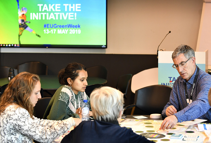 Mensen discussiëren rond een tafel vol documenten, voor een scherm met informatie over de Groene Week