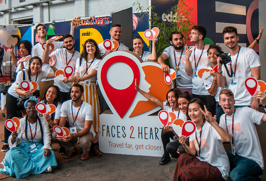 Een groep jongeren verzameld rond een kartonnen bord met het logo van “Faces2Hearts”