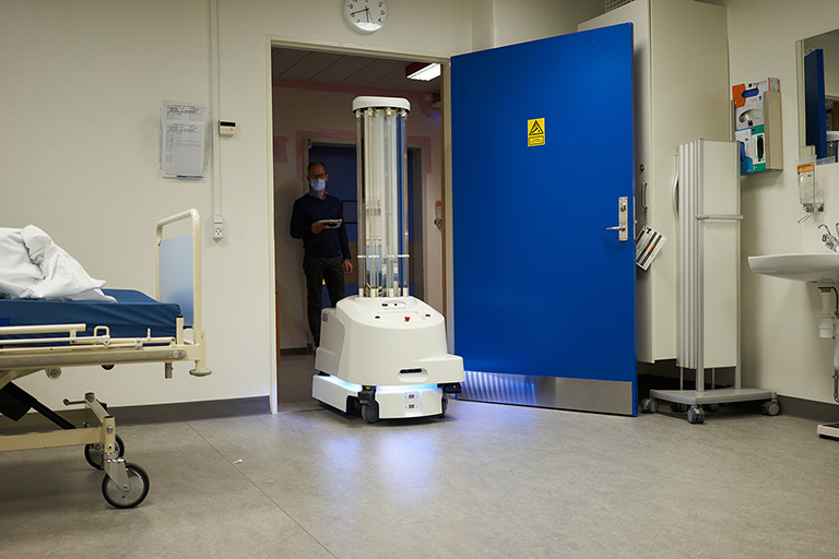 A machine rolling in to a hospital room remotely while a healthcare worker controls it from the background.