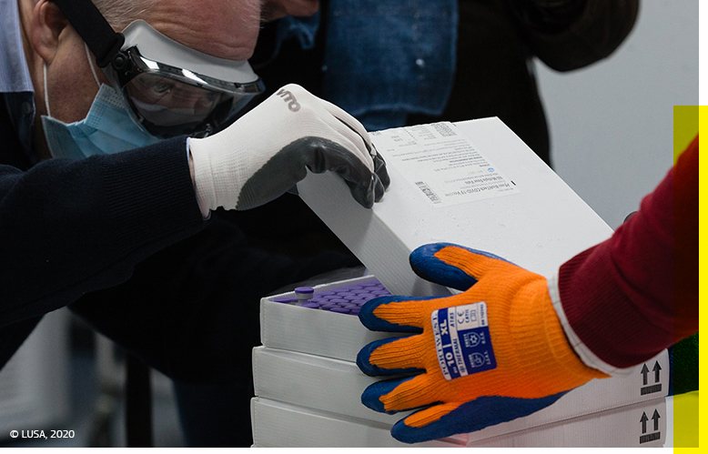 A close-up of a man in protective wear opening the top of a stack of boxes containing medical vials while supervised by a colleague in protective wear. © Lusa 2020.