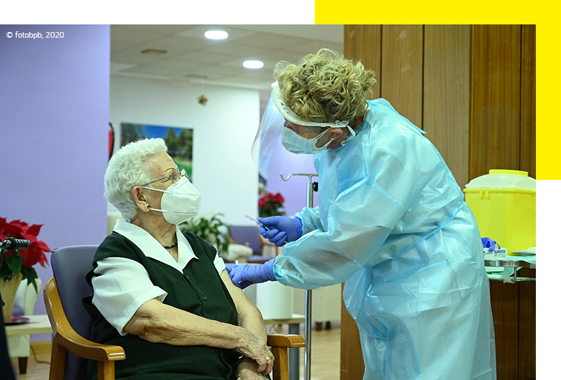 Een oudere vrouw met mondmasker wordt gevaccineerd door een zorgverlener in beschermende kleding. © fotobpb, 2020