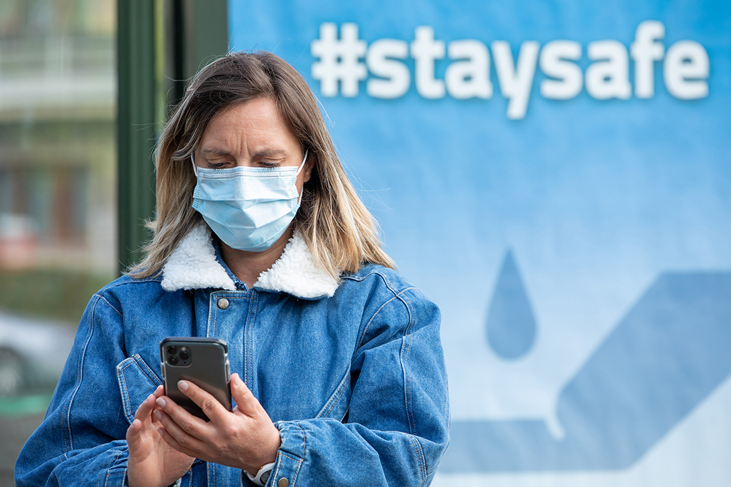 En kvinna iförd munskydd står i förgrunden och tittar på sin telefon framför en utomhuskampanj med covid-19-information.