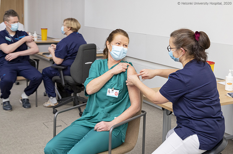 A medical worker in scrubs and a mask receives a vaccine from a colleague, with other medical workers being vaccinated in the background. © Helsinki University Hospital, 2020.