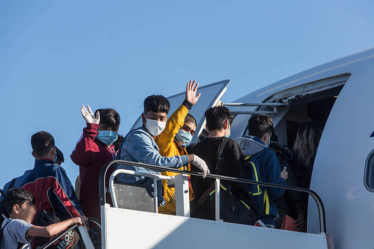 A group of teens and children wearing masks, waving while boarding a plane.