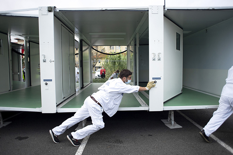 A man in white scrubs pushes interlocking makeshift medical-examination rooms across a car park.