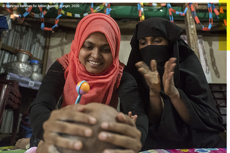 Two Rohingya women with a toy rattle clap to amuse a child. © Mahmud Rahman for Action Against Hunger, 2020.