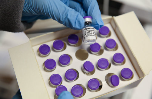 Close-up van een in een medische handschoen gestoken hand die één flacon uit een doos vaccins pakt.
