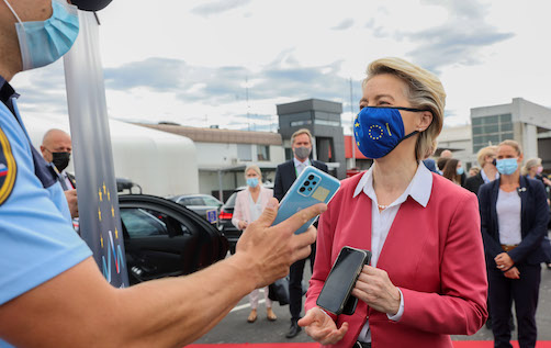 Ursula von der Leyen wears a mask while a masked officer scans her phone.