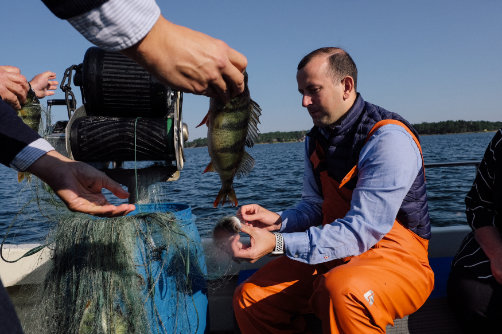 Virginijus Sinkevičius egy hajón, halászeszközt a kezében tartva, miközben egy halász felmutatja a fogást.