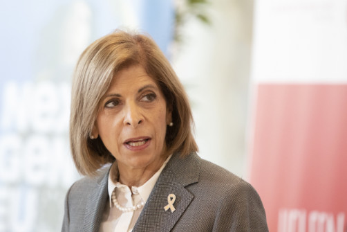Stella Kyriakides wearing a breast-cancer pin and looking beyond the camera.