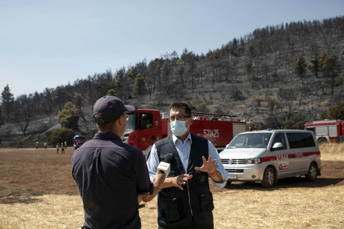 Janez Lenarčič speaking to a reporter with a forest and emergency vehicles in the background.