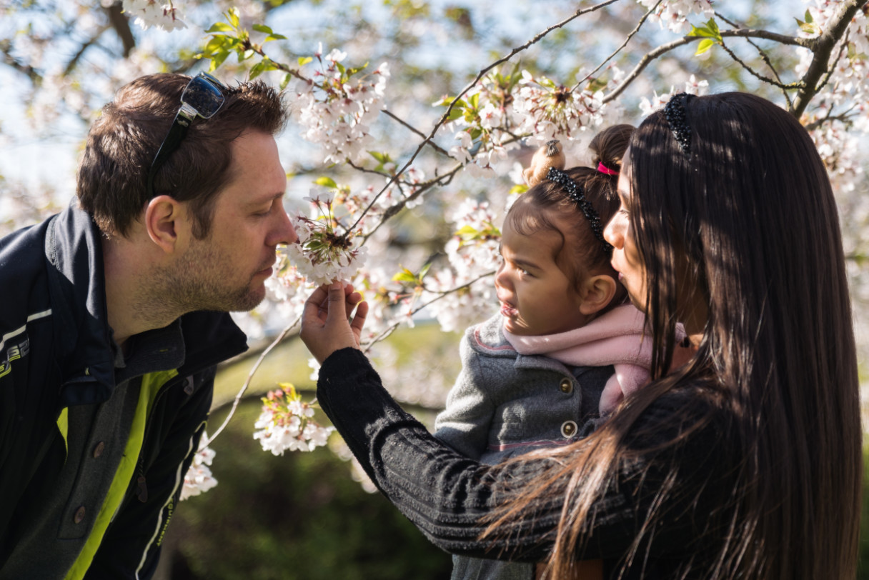 Egy kisgyermekes fiatal család a parkban, amint egyikük egy virágot szagol meg.