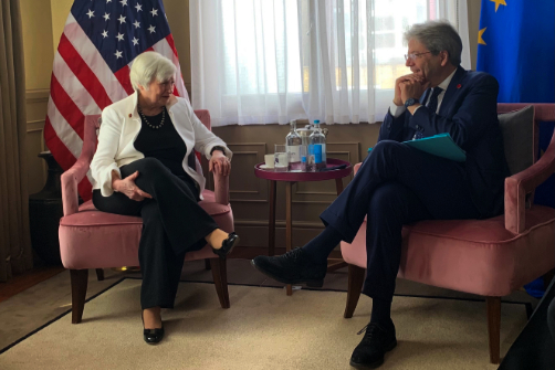 Paolo Gentiloni charla con Janet Yellen, ambos sentados delante de las banderas de la Unión Europea y de los Estados Unidos, respectivamente.