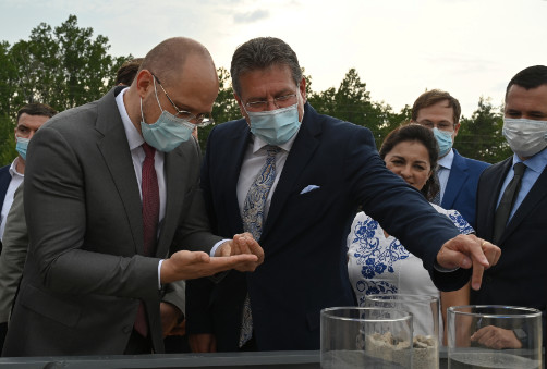Maroš Šefčovič a Denys Shmyhal v rouškách si prohlížejí vzorky, v pozadí menší skupina lidí.