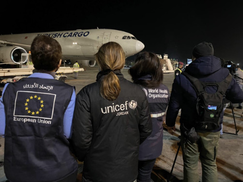 Няколко души, облечени в якета с емблемата на Европейския съюз, УНИЦЕФ или Световната здравна организация на гърба, гледат към самолет на пистата. Запазени права — Световна здравна организация, две хиляди двадесет и втора година.