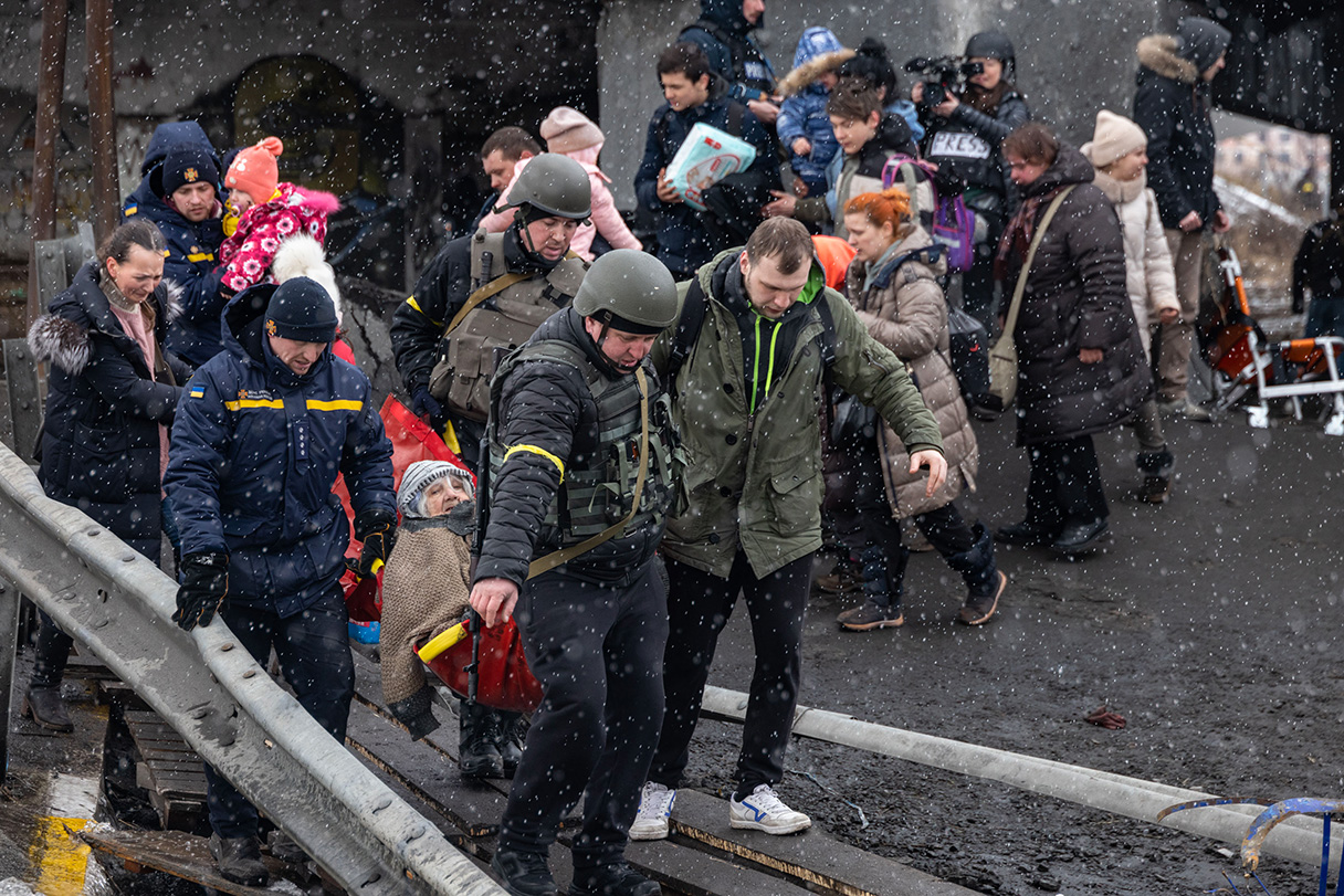 Grupa Ukraińców przechodzi prowizoryczną kładką, czterech mężczyzn niesie na noszach starszą kobietę.