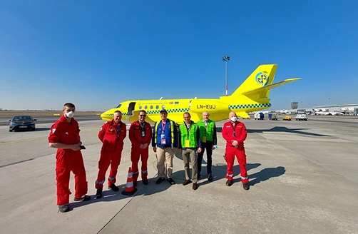 Septyni medicininę evakuaciją vykdantys darbuotojai stovi aerodrome, už jų – medicininės evakuacijos lėktuvas.