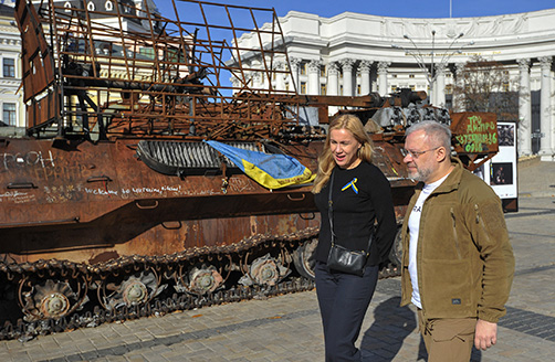 Kadri Simson, wearing a ribbon with the Ukrainian flag pinned to her shirt, and Herman Haluschenko walk past a damaged tank in the background while talking to each other.