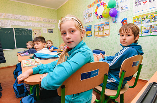 Cinci copii ucraineni stând în bănci într-o clasă, întorși cu fața spre cameră.