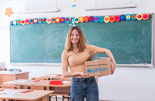 Anastasiia Konovalova står leende i ett klassrum med en Unicef-låda under armen.
