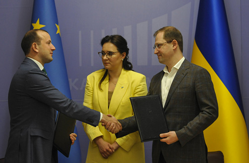 Virginijus Sinkevičius and Ruslan Strilets shake hands in front of a European flag and a Ukrainian flag, while Yulia Svyrydenko stands between them, slightly to the rear, facing Virginijus Sinkevičius.