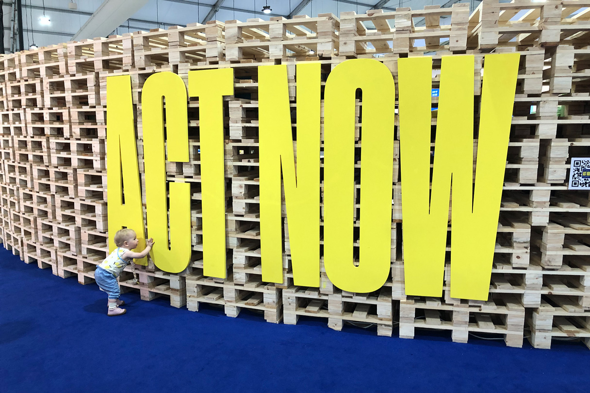 Letters forming the words ‘ACT NOW’ mounted on stacked pallets with a toddler touching them.