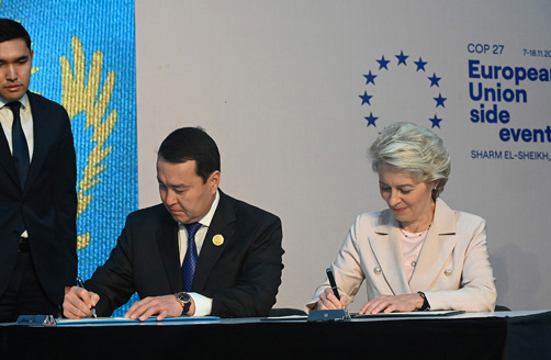 Alikhan Smailov e Ursula von der Leyen sentados à mesa lado a lado assinam documentos.