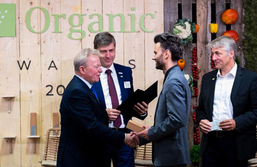 Janusz Wojciechowski îi strânge mâna unui câștigător în fața unui perete din lemn cu inscripția I U Orghenic ăuards 2022.