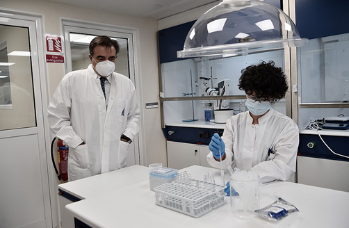 Margaritis Schinas, som bär laboratorierock och munskydd, observerar en laboratorietekniker.