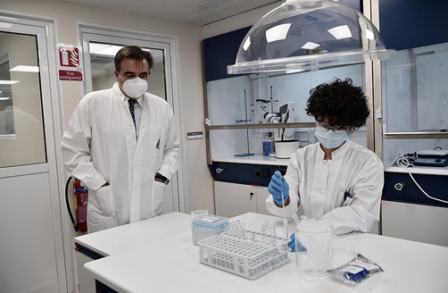 Margaritis Schinas u laboratorijskom ogrtaču i s maskom na licu promatra laboratorijsku tehničarku pri pipetiranju.