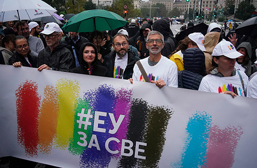 Helena Dalli loopt mee in de Pride-optocht, achter een spandoek met de hashtag “EU voor iedereen” in Servisch Cyrillisch schrift op een achtergrond met regenboog.