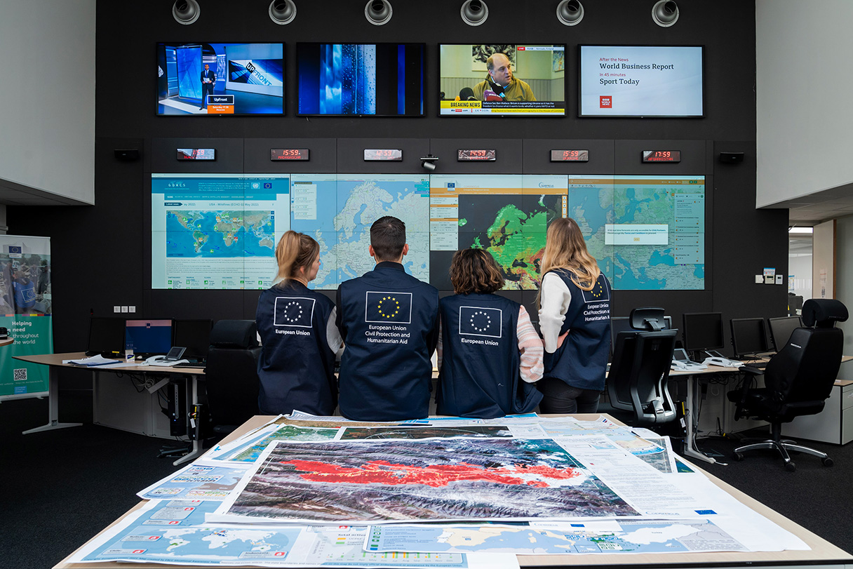 Fyra av EU:s civilskydds- och biståndsarbetare i arbetskläder på ett kontor, som tittar på en stor skärm med flera kartor.