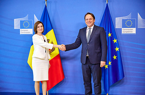 Maja Sandu ir Oliveris Varhėjis Moldovos ir Europos Sąjungos vėliavų fone žiūrėdami į fotoaparato objektyvą spaudžia vienas kitam ranką.