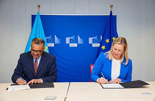 Tedros Adhanom Ghebreyesus och Jutta Urpilainen undertecknar ett avtal vid ett bord med Världshälsoorganisationens flagga, en EU-flagga och en blå banner med tre logotyper för Europeiska kommissionen bakom dem.