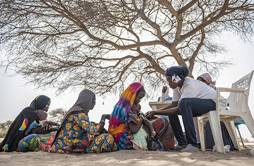 Tre kvinnor sitter i rad på marken, i skuggan av ett träd, i flyktinglägret Kousseri i Tchad, med sina barn i knät. En barnmorska sitter vänd mot dem och tittar på det första barnet i raden.