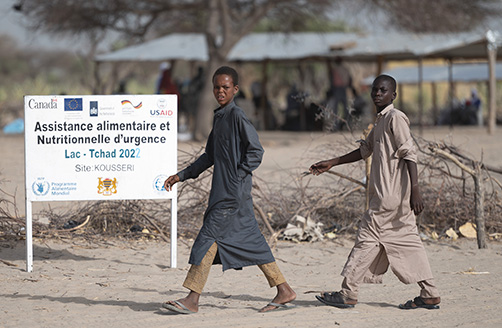 Doi tineri trec pe lângă un panou în limba franceză pe care scrie „Ajutor alimentar de urgență” în tabăra de refugiați Kousseri din Ciad.
