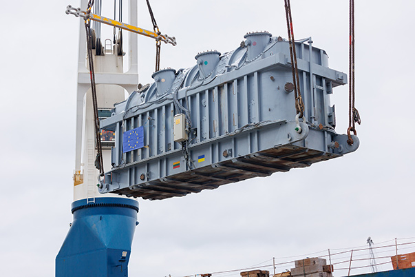 Ein Großstromgenerator hängt an einem Kran und wird durch die Luft geschwenkt und auf ein Schiff verladen.