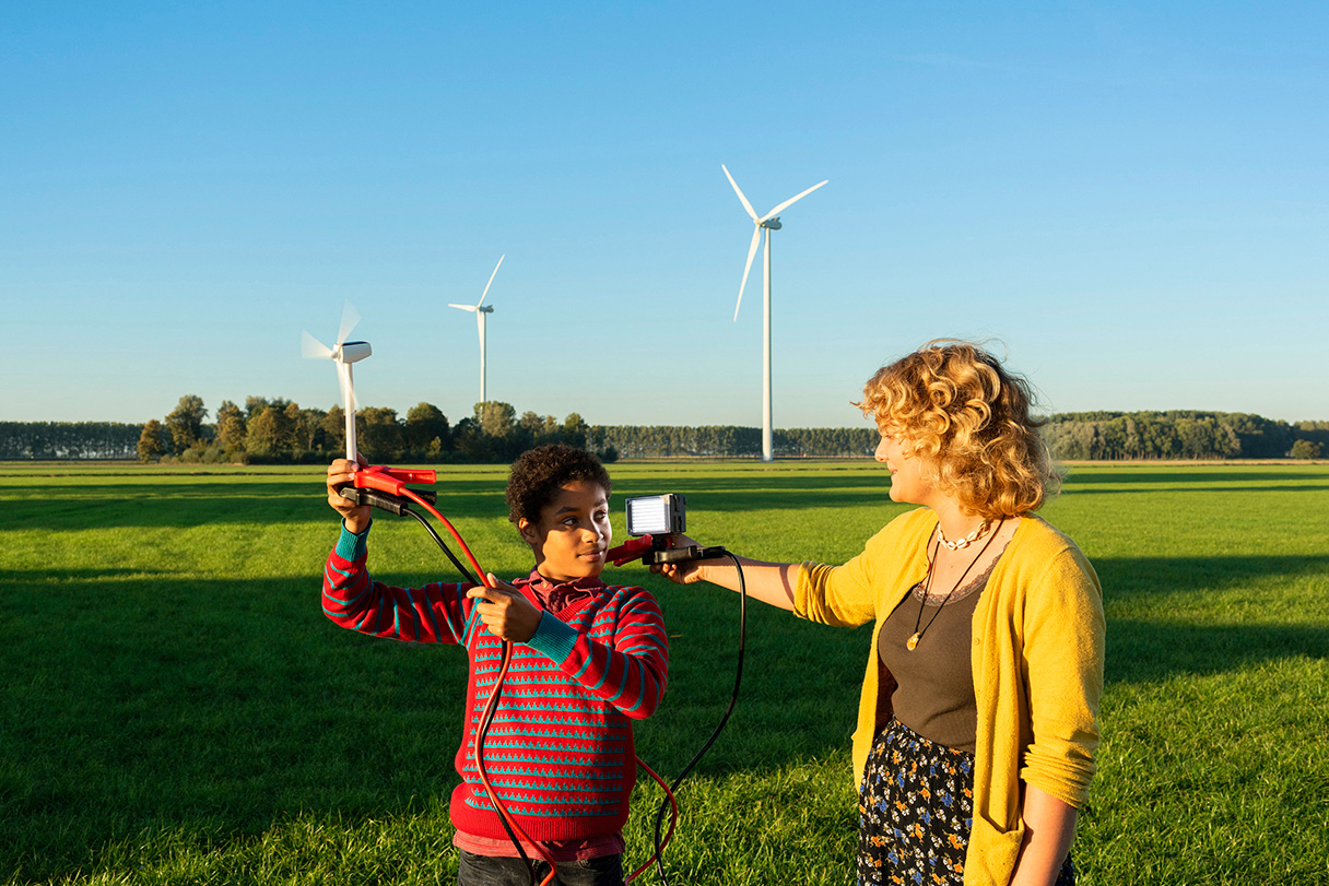 Dos jóvenes están de pie en un campo cubierto de hierba con dos turbinas eólicas visibles detrás de ellos. Uno sostiene un modelo de turbina eólica en miniatura conectado a un dispositivo portátil que sujeta el otro joven con la mano.