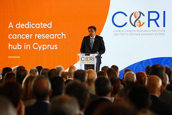 Margaritis Schinas steht hinter einem Rednerpult auf einer Bühne vor einem großen Publikum. Auf dem Bildschirm hinter ihm werden die Worte „A dedicated cancer research hub in Cyprus“ angezeigt.