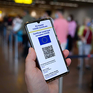 Eine Hand hält ein Telefon, das einen QR-Code und die Worte „EU digital COVID certificate“ anzeigt. Im Hintergrund stehen Menschen mit Koffern Schlange.